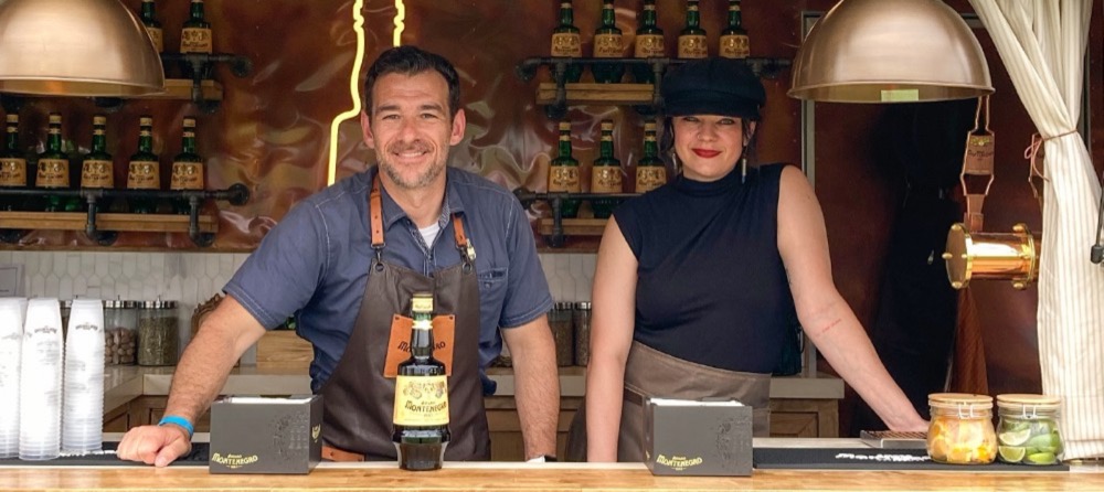 Two bartenders in leather aprons smile across the counter of the Bar Monte trailer, a bottle of Amaro Montenegro between them, with copper walls, a neon bottle outline, iron pipe shelves of Montenegro bottles, copper pendant lamps and jars of citrus garnish behind and beside them.