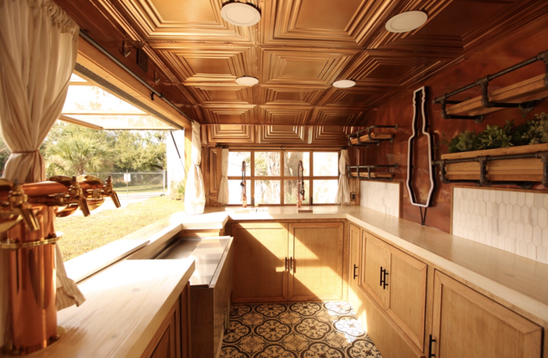 Inside the Bar Monte trailer: a copper-toned pressed ceiling, wood cabinets under pale counters, a copper draft tower at the open service window, herb planters on black iron pipe shelves, a bottle-shaped outline sign and a patterned tile floor.