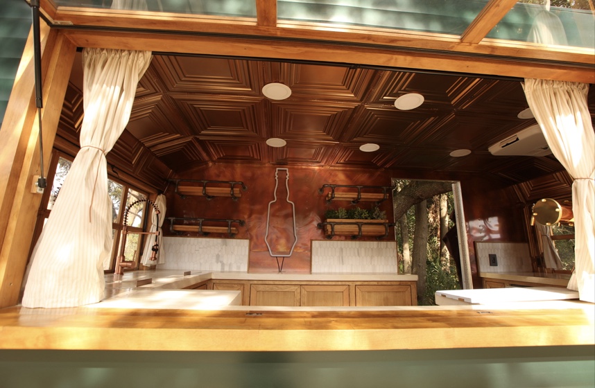 View through the open service window of the Bar Monte trailer, framed by cream curtains, showing a wooden counter, the copper ceiling, pipe shelving and a bottle-shaped outline on the back wall.