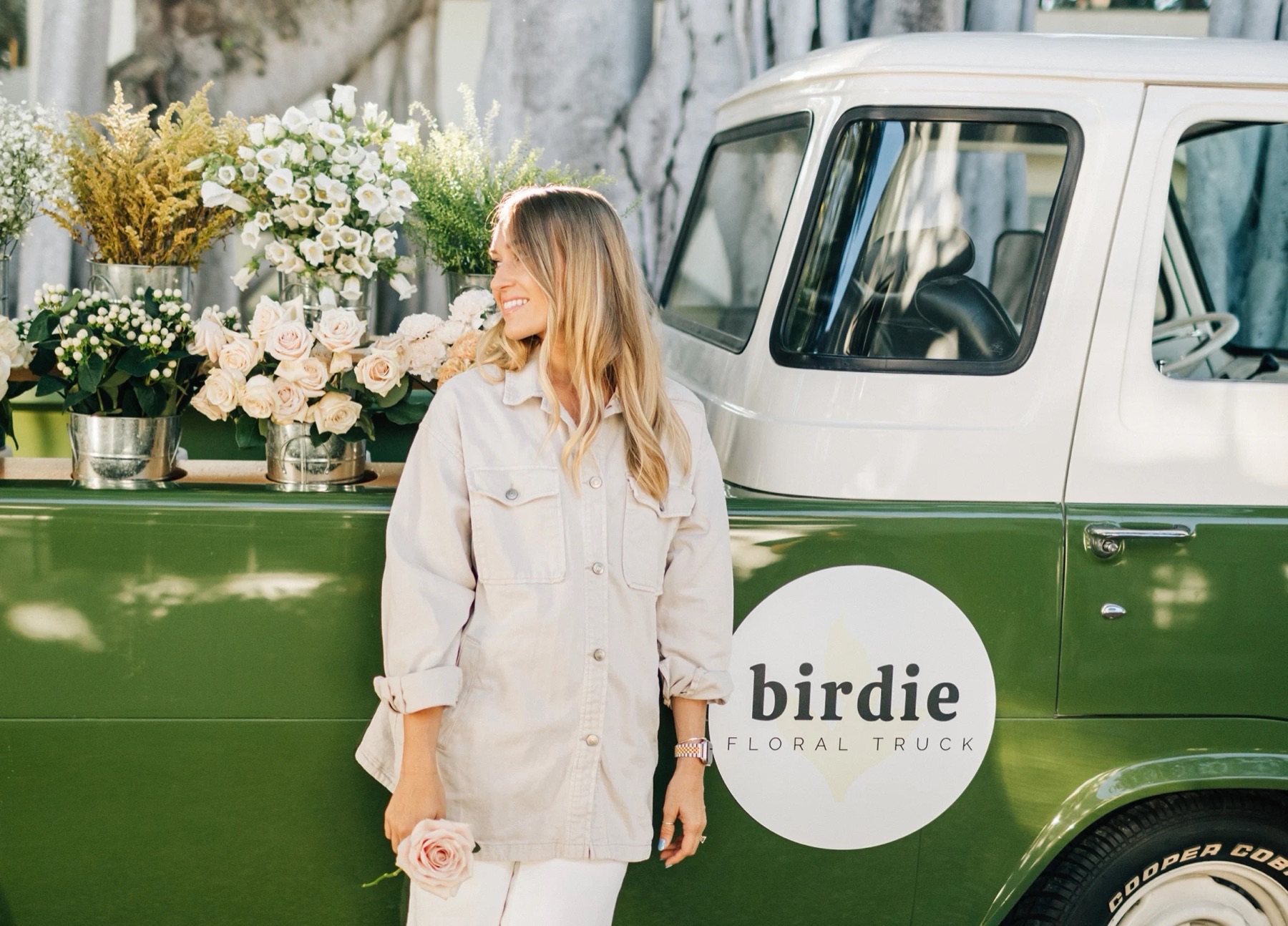 A woman stands beside a green-and-white vintage pickup with a round "birdie floral truck" logo on its door and buckets of white and pale pink flowers along its bed.
