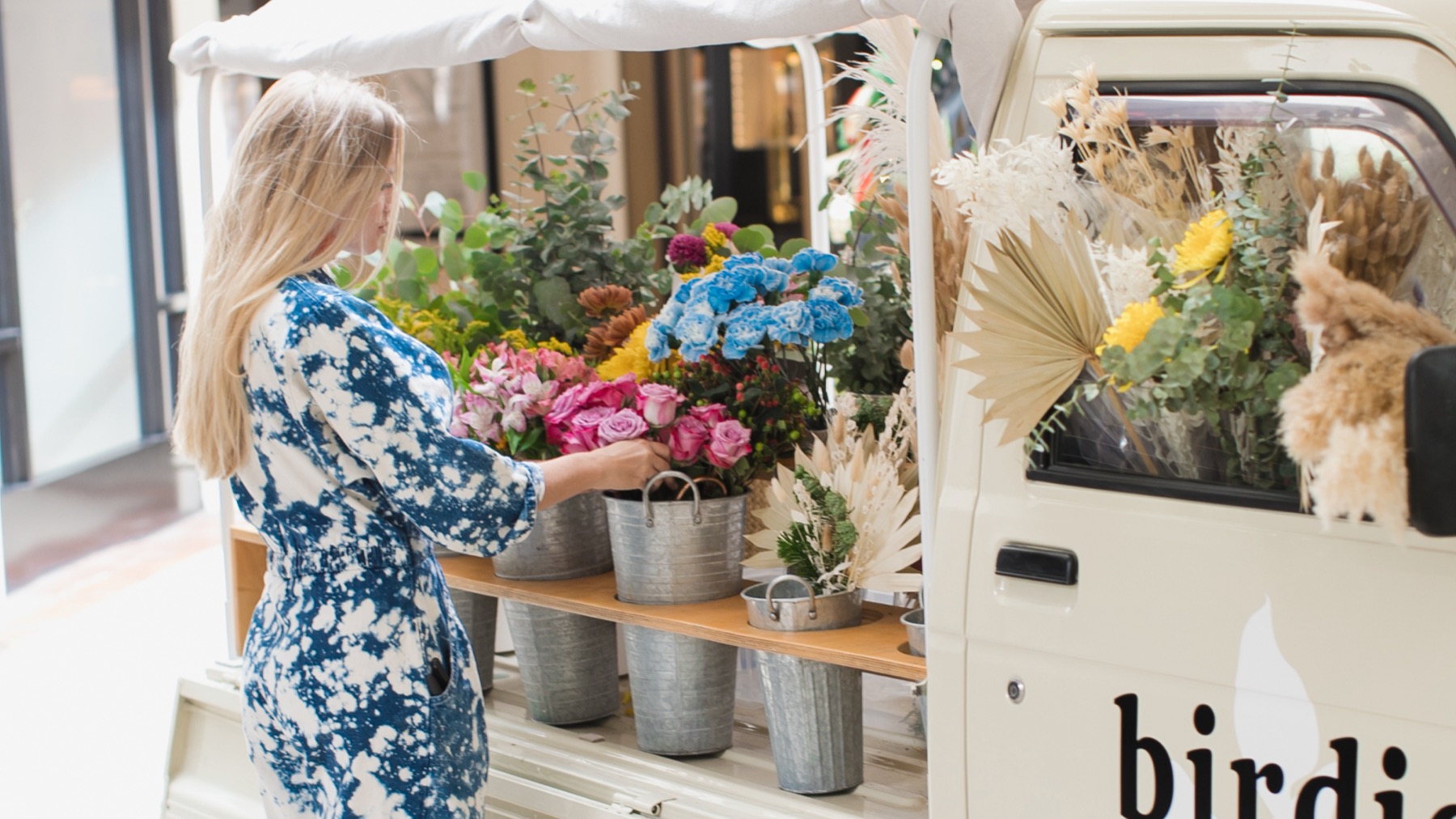 A woman arranges pink roses in galvanized buckets on the wooden shelf of the Kei truck's canopied bed, with dried palm and pampas stems tucked against the cab window.