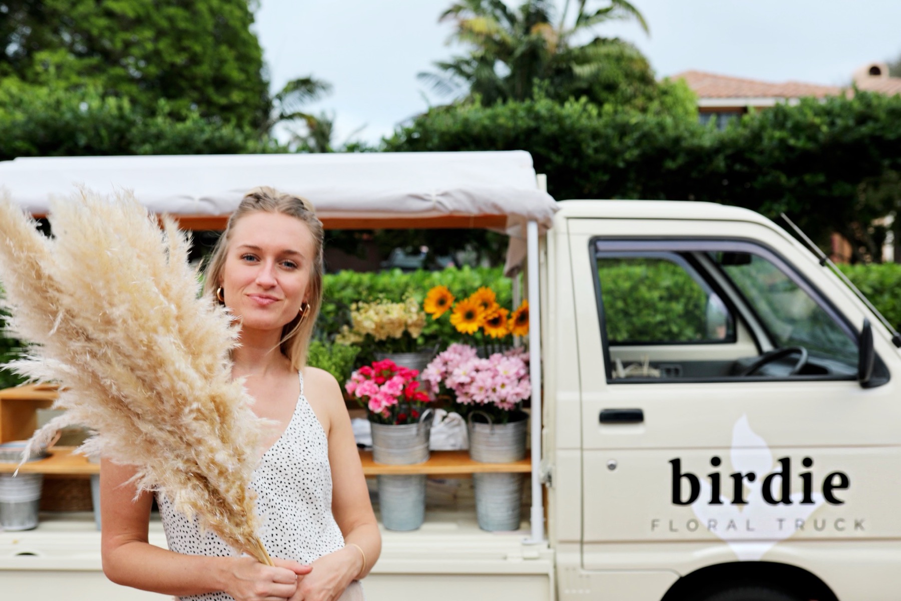 A woman holding a large bundle of pampas grass stands beside a small cream-colored Kei truck branded "birdie floral truck," its canopied bed filled with buckets of flowers.