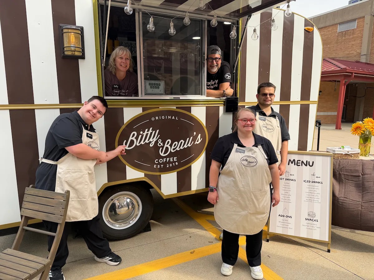 Four Bitty & Beau's employees in aprons stand beside a brown-and-white striped Coffee Cruiser, one pointing at the oval logo, while two more people smile from the open service window; a sandwich-board menu stands at right.
