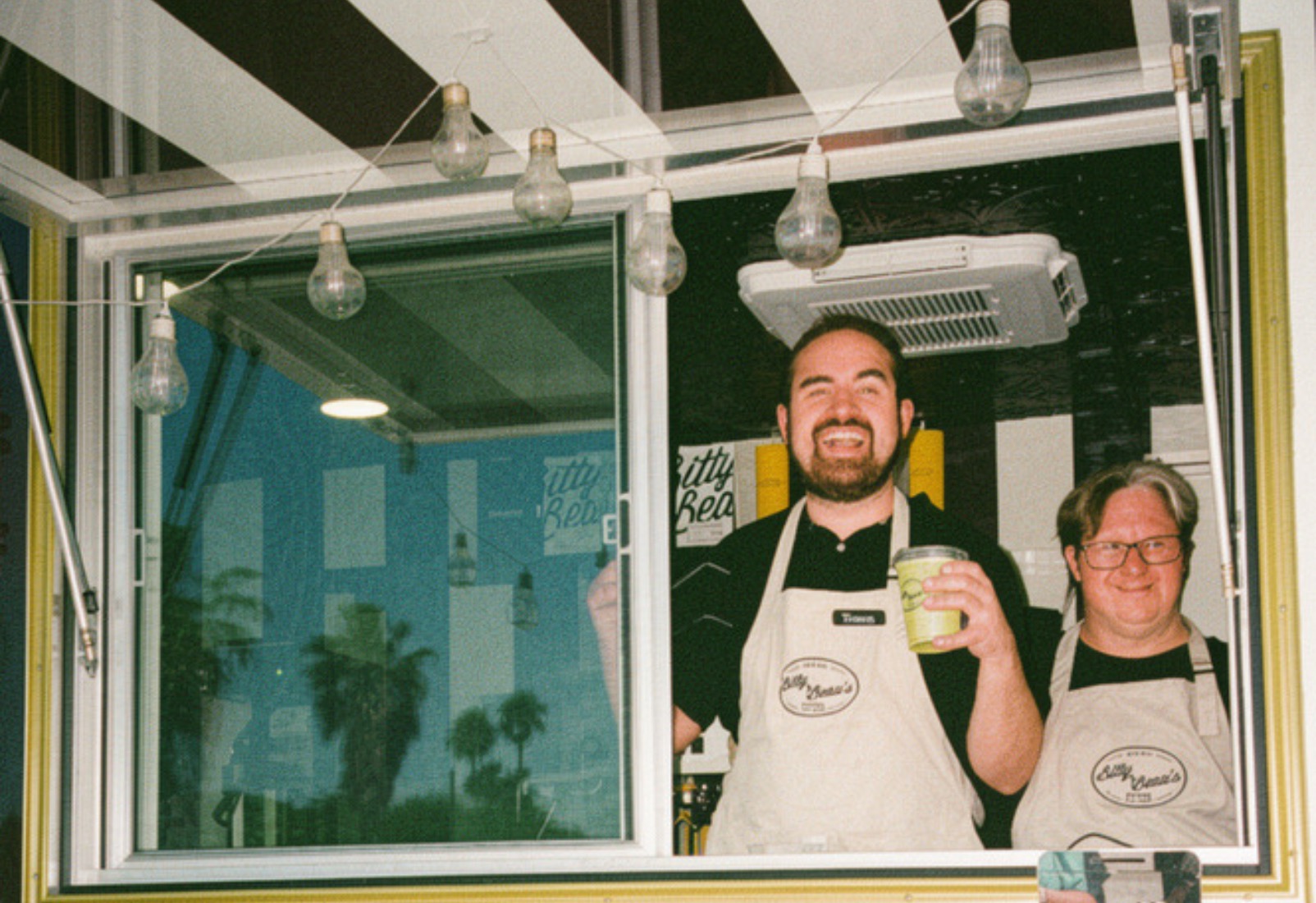 Two smiling Bitty & Beau's employees in aprons look out from a Coffee Cruiser's open service window under strung bulbs, one raising a cup.