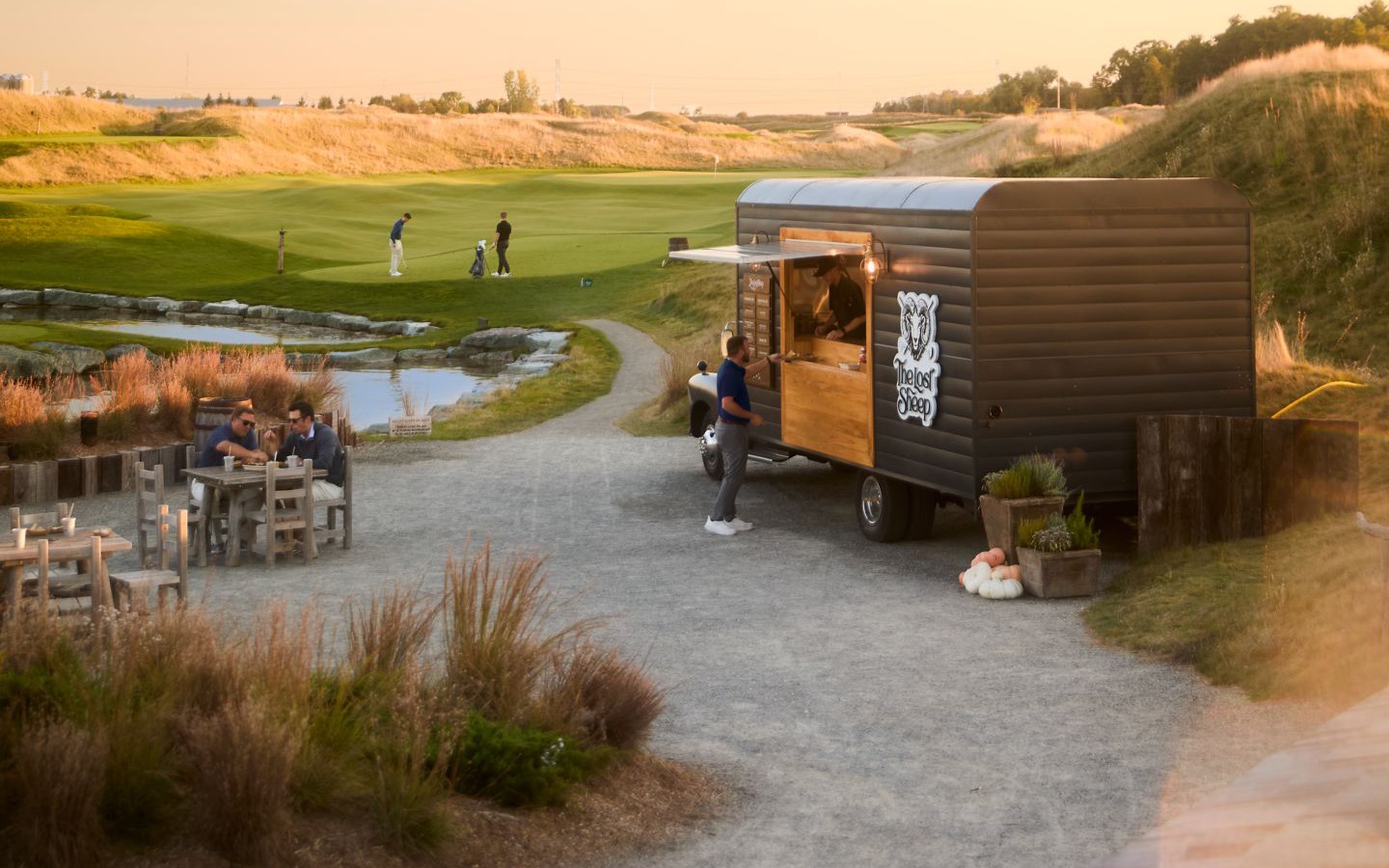 A dark food truck with a wood-framed serving window and a ram's-head Lost Sheep sign parked on a gravel apron at golden hour, with two men eating at a wooden table to the left and golfers on a green beyond a stone-edged pond.