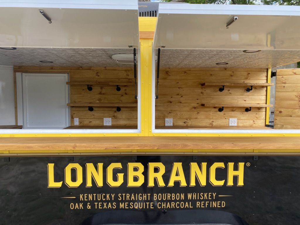 Two raised service hatches on the Longbranch trailer opening onto a pine-paneled interior with wooden shelves on black pipe brackets, above a wooden counter and the words Longbranch Kentucky Straight Bourbon Whiskey, Oak and Texas Mesquite Charcoal Refined.