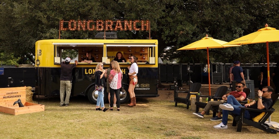 The yellow-and-black Longbranch horse trailer serving at an outdoor festival under a lit LONGBRANCH marquee sign, two bartenders at its open service windows and guests at the counter, with yellow umbrellas and Adirondack chairs to the right and a wooden Longbranch Humanshoes game box on the grass at left.
