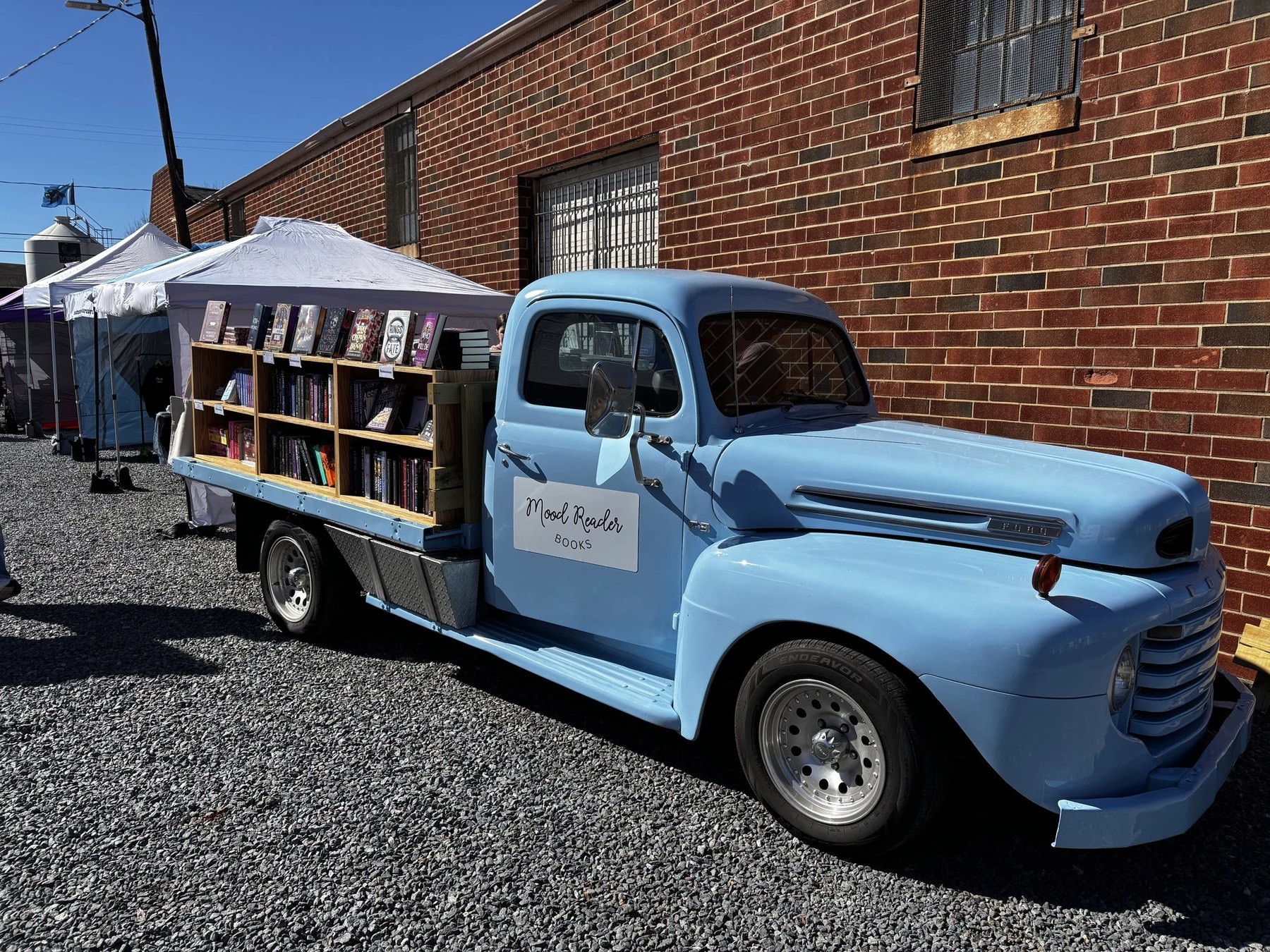 A restored light blue Ford pickup with a 'Mood Reader Books' sign on the door, its flatbed fitted with wooden shelves of books, parked on gravel beside a brick building with market tents behind.