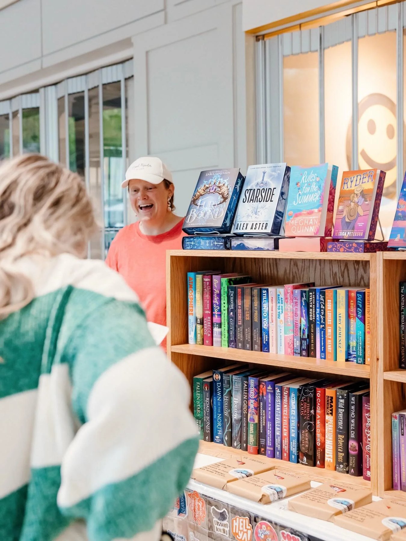 A shopper in a green and white sweater faces a wooden bookcase of romance and fantasy titles, with face-out new releases on top, as a bookseller in a Mood Reader Books cap smiles behind it.