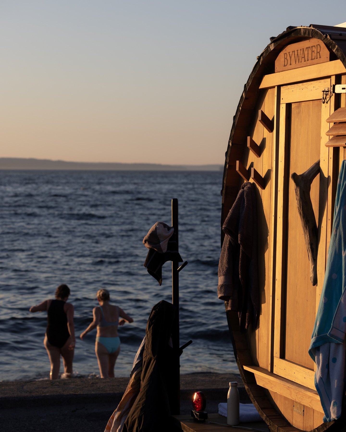 The end of a wooden barrel sauna carved with the name Bywater, towels hanging on its hooks, as two bathers wade into the water at dusk.