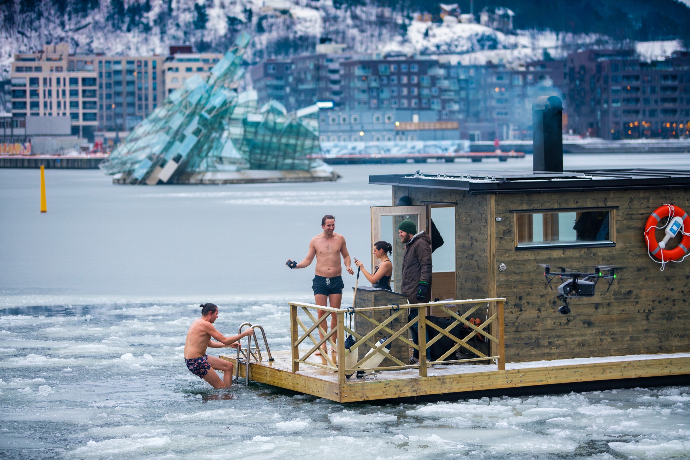 Bathers on the deck of a small wooden floating sauna in an icy Oslo harbor, one climbing down a ladder into the water, with the glass sculpture She Lies and waterfront apartments behind.
