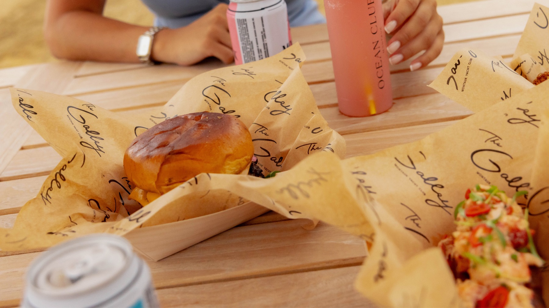 A burger and a lobster roll served in paper boats lined with paper printed in "The Galley" script, on a wooden table with canned drinks.