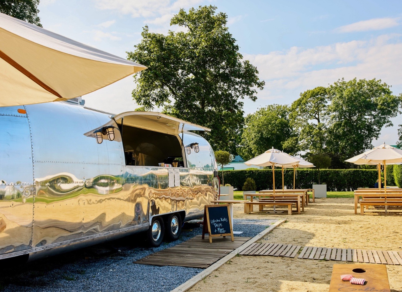 A polished aluminum Airstream trailer parked on gravel beside a wooden walkway, its serving hatch propped open under two caged lamps and a chalkboard sign at its side, with picnic tables and white umbrellas set on sand and a lawn and trees behind.