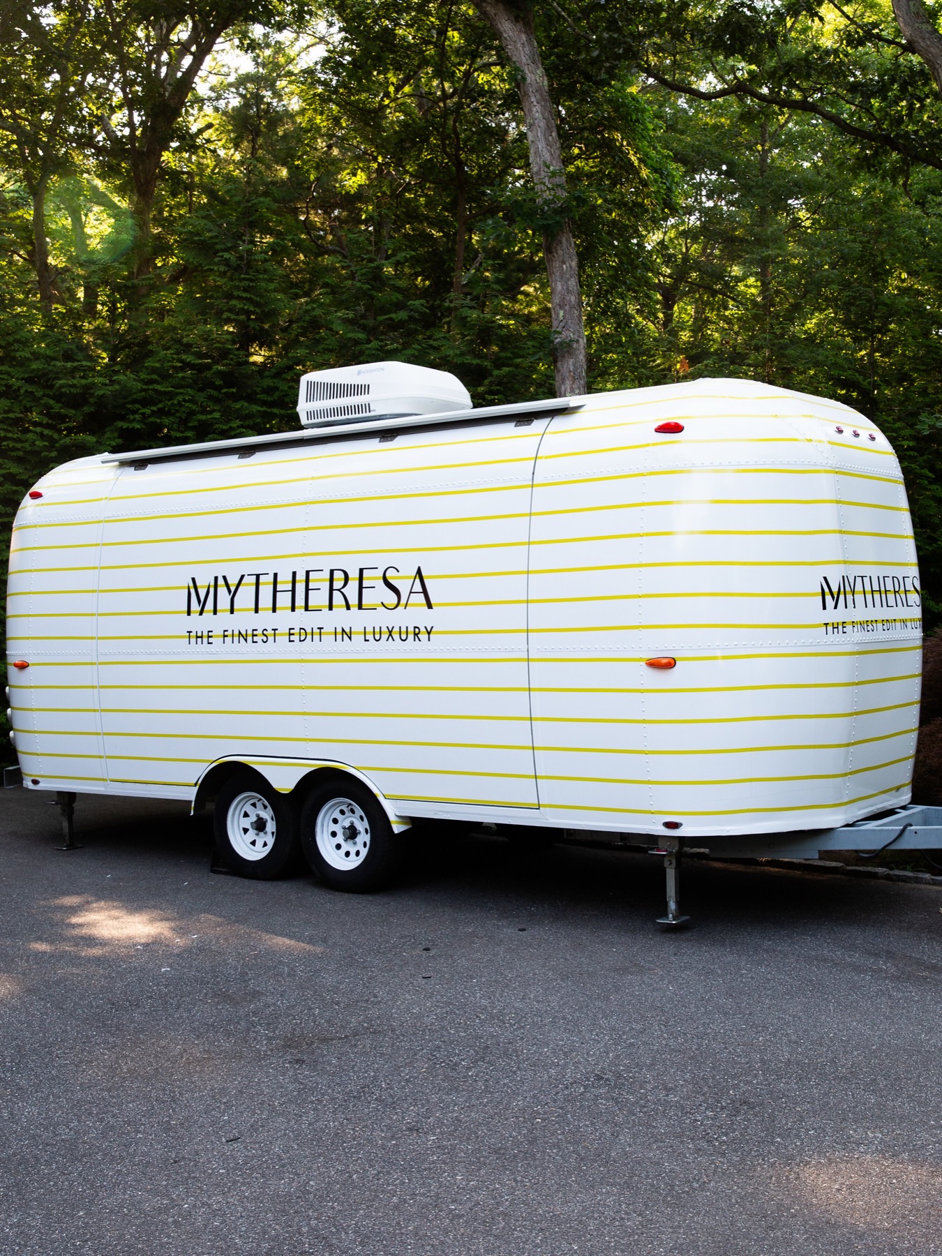 The closed Airstream trailer in white with thin yellow stripes and the Mytheresa wordmark and "The Finest Edit in Luxury" on its side, parked on a driveway under trees.