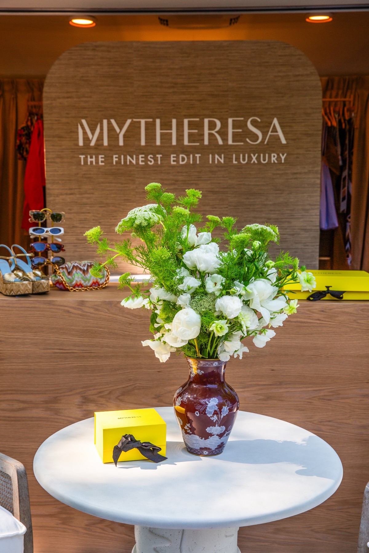 Inside the trailer: a woven wall panel with the Mytheresa logo above a wooden counter displaying sandals, with a vase of white flowers on a round white table in front.