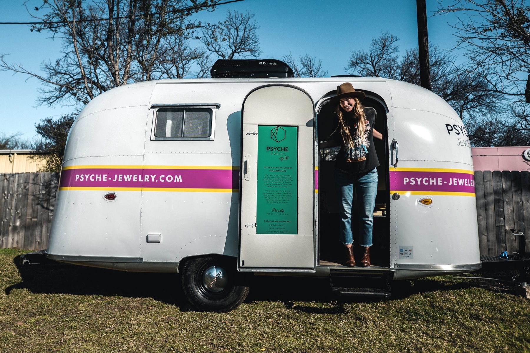 A small white vintage Airstream trailer with a magenta and yellow stripe reading "psyche-jewelry.com," parked on grass, with a woman in a wide-brimmed hat standing in the open doorway.