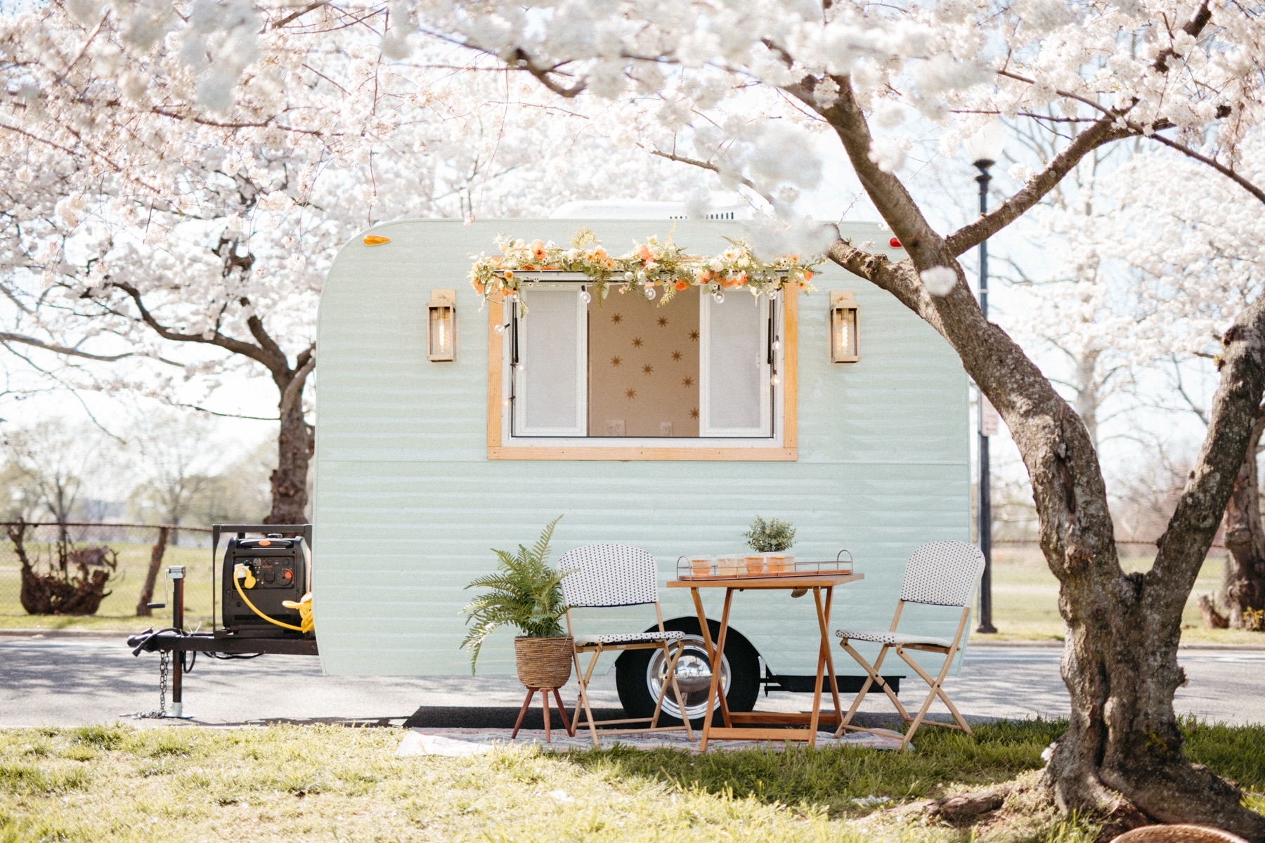A mint-green vintage-style camper trailer with a flower garland over its wood-framed service window and brass wall lanterns, parked under white blossoming trees with a bistro table, two folding chairs and a portable generator on the tongue.