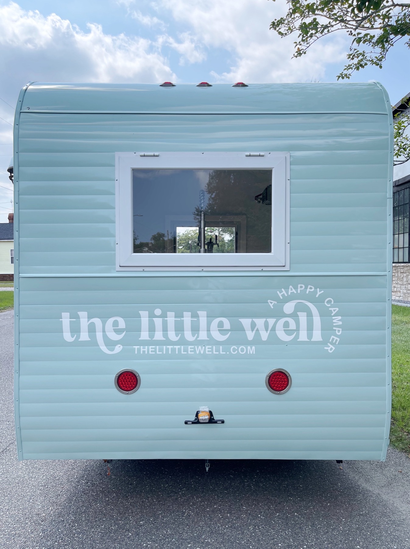 Rear view of the mint-green camper trailer with a small window, two round red tail lights and white lettering reading 'the little well, a happy camper, thelittlewell.com'.
