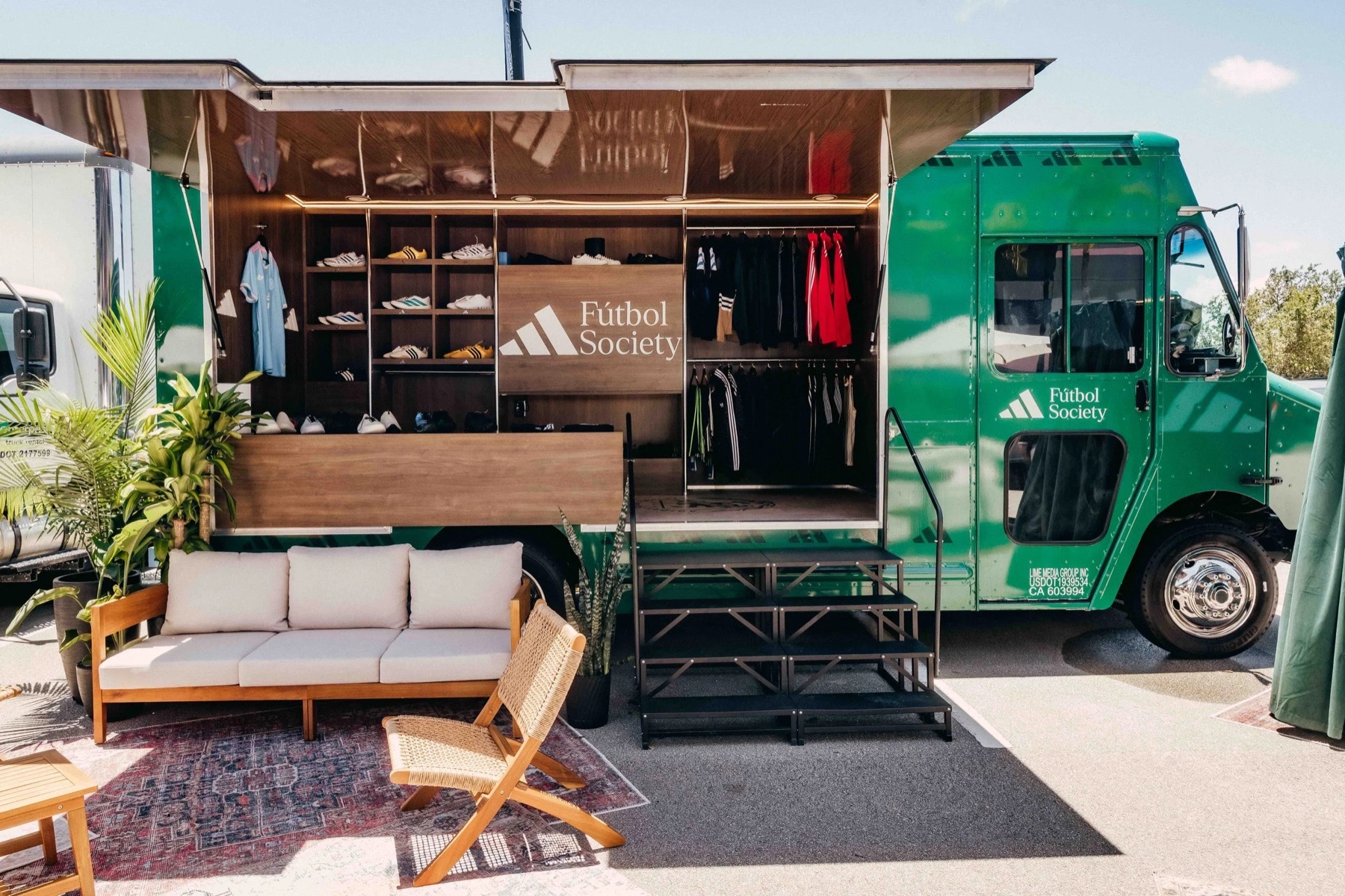 A green Adidas Fútbol Society truck with its side opened to show wood-paneled shelving of sneakers and jerseys, a sofa, chair and rug set out in front.
