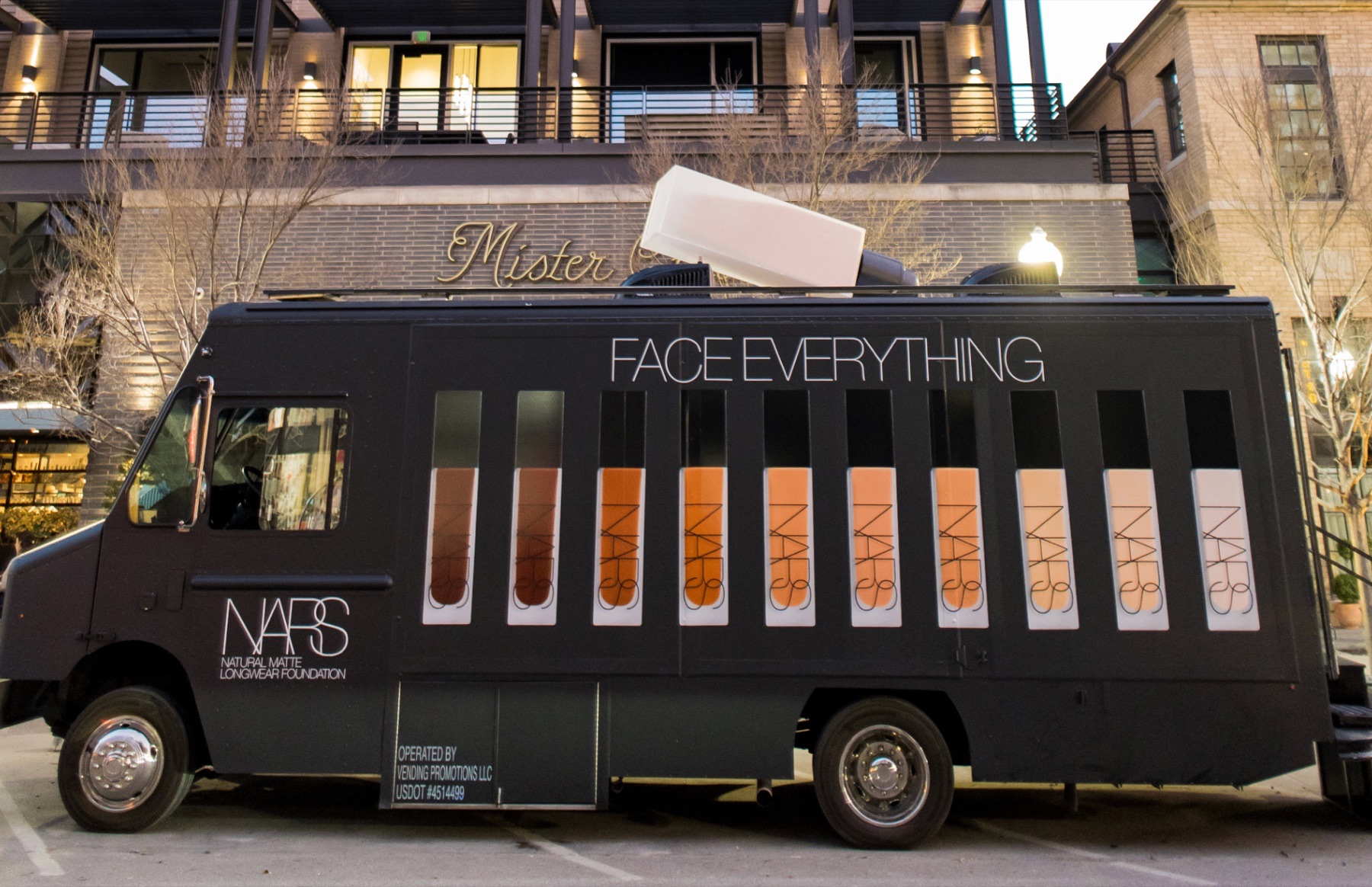 A black box truck reading 'Face Everything' with rows of NARS foundation bottles in its side windows and an oversized bottle on the roof, parked outside a building at dusk.