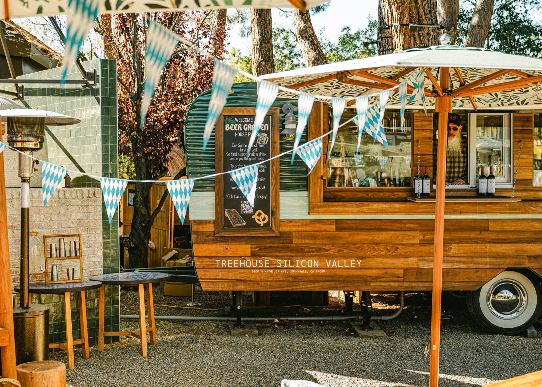 The green-and-timber beer trailer in service in the Treehouse Silicon Valley Beer Garden, under blue-and-white Bavarian bunting and a leaf-print umbrella, with a bearded bartender at the open service window, wine bottles on the counter, a chalkboard of Beer Garden house rules on its side and "Treehouse Silicon Valley" lettered on the timber cladding.