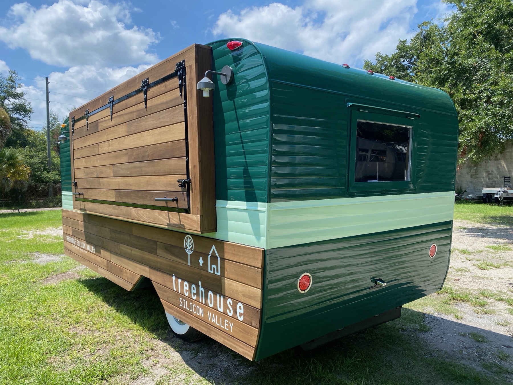 The green-and-timber trailer from the rear corner with its service side shut behind a hinged timber panel held by black strap hinges and latches.