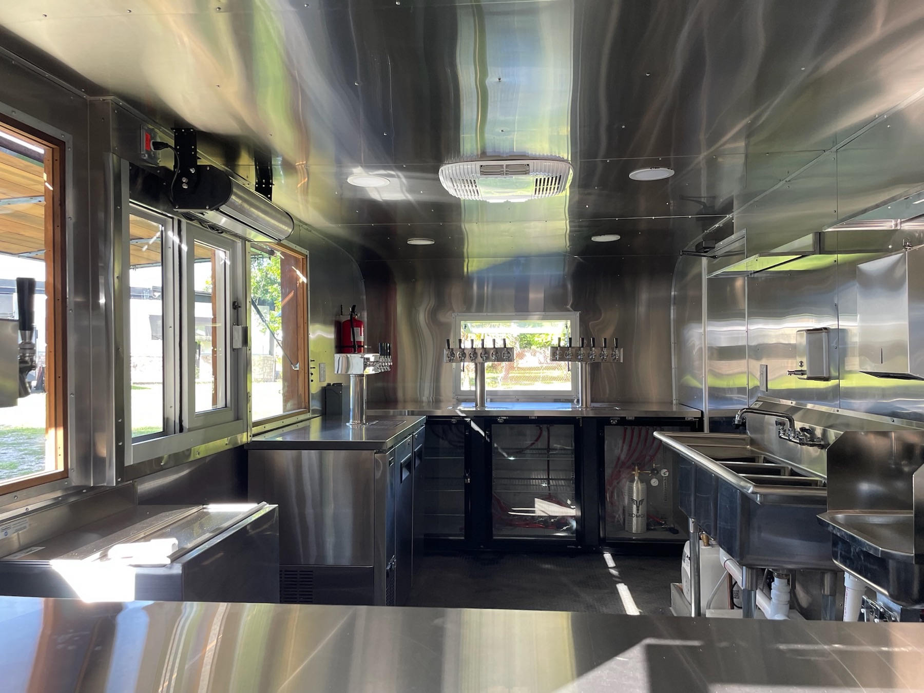 The trailer's interior lined in polished stainless steel, with two multi-tap beer towers over glass-door refrigerators, a separate tap tower by the service windows, and a three-compartment sink and hand sink on the right.