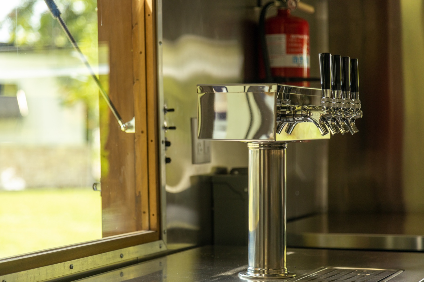 A chrome T-shaped beer tower with a row of black-handled faucets on a stainless counter beside the trailer's open service window.